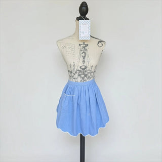 Her Good Eye cornflower blue half apron with a gathered waist, side pocket, and white scalloped hem on a mannequin.