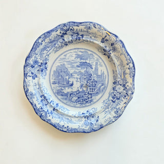 Her Good Eye blue and white ceramic dinner plate with a scalloped edge and intricate pastoral transferware scene.