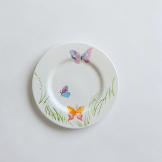 Her Good Eye white ceramic decorative plate with hand-painted butterflies in pink, blue, orange, and purple, and green grass accents.