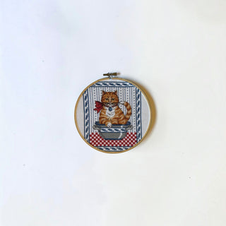 Her Good Eye vintage needlepoint art featuring an orange tabby cat in a bowl, framed in a round wooden embroidery hoop.