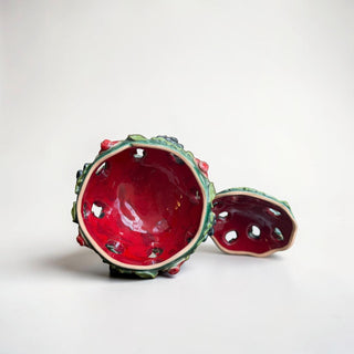 Her Good Eye decorative ceramic jar with emerald green glaze, sculpted fruits, and leaves. Red apples and grapes adorn the exterior. The lid is open, revealing a glossy red interior.