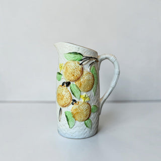 Her Good Eye ceramic pitcher with embossed golden pears, green leaves, and a bamboo-style handle on a woven texture.