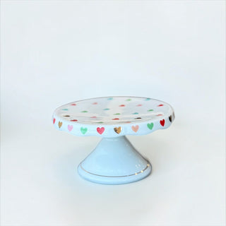 Her Good Eye ceramic pedestal cake stand in pastel blue with a colorful heart pattern and glossy finish.