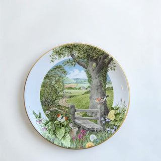 Her Good Eye decorative plate with a pastoral scene: oak tree, woodpecker, finch, rabbit, butterflies, wildflowers, cottages, and cattle. Gold rim.