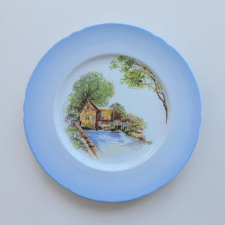 Her Good Eye decorative ceramic plate with landscape scene, cottage, river, bridge, and scalloped edge. Sky blue rim.