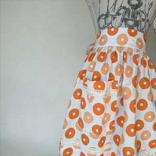 Her Good Eye donut print cotton apron with pockets and lace trim. Orange and peach donuts on white.