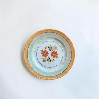 Her Good Eye decorative plate with scalloped gold rim, pale green iridescent background, and floral motif with orange and blue flowers.