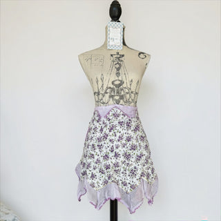 Her Good Eye floral print ruffled half apron with purple wildflowers on white and lavender trim. Features a pocket.