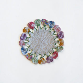 Her Good Eye handmade crochet doily with a neutral center and a border of multicolored pansy flowers.