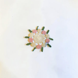 Her Good Eye handmade crochet doily with pink and cream floral roses and green leaves on a white background.