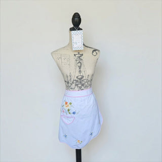 Her Good Eye vintage style white half apron with floral embroidery, pink piping, and a front pocket on a mannequin.