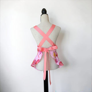 Her Good Eye light pink floral print cotton blend apron with coral ruffles, criss-cross straps, and bow accent.
