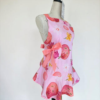 Her Good Eye light pink floral print cotton blend apron with ruffled hem and coral bows. Features daisies and abstract circular motifs.