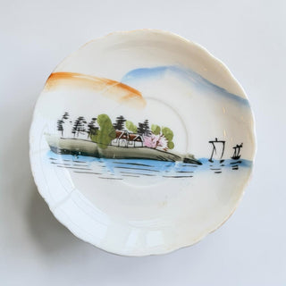Her Good Eye porcelain saucer featuring a hand-painted coastal landscape with a cottage, trees, and sailing vessels.