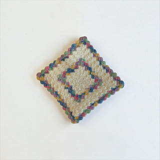 Her Good Eye handmade square crochet drink coaster in cream cotton with colorful blue, pink, green, and gold accents.