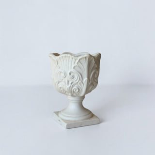 Her Good Eye matte white ceramic pedestal urn with ornate scrollwork and scalloped rim on a square base.