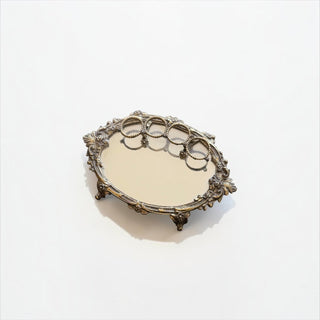 Her Good Eye oval vanity mirror tray with an ornate antique-gold floral frame and decorative feet on white background.