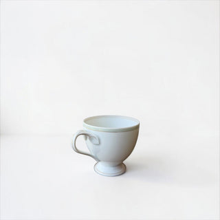 Her Good Eye white porcelain tea cup with a pedestal base, ergonomic handle, and subtle rim detail on a white surface.