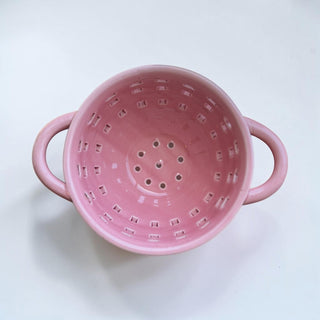 Her Good Eye pink ceramic colander with handles and cutout pattern, perfect for draining pasta or rinsing produce.