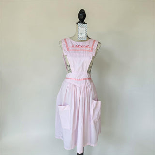 Her Good Eye light pink cotton blend pinafore apron with "SINGER" embroidery, ribbon trim, adjustable straps, tie waist, and two front pockets.