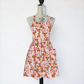 Her Good Eye strawberry print cotton apron with adjustable halter straps and two front pockets. Features pink strawberries and brown leaves on white.