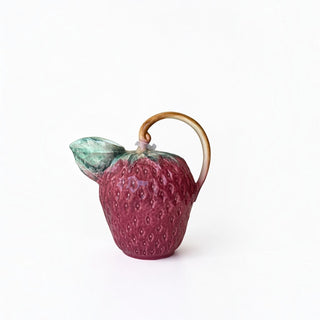 Her Good Eye strawberry-shaped ceramic pitcher with a glossy red textured body, green leaf spout, and vine handle.