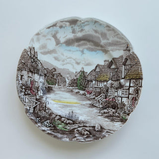 Her Good Eye decorative ceramic plate featuring a detailed village scene with thatched-roof cottages and a winding lane.