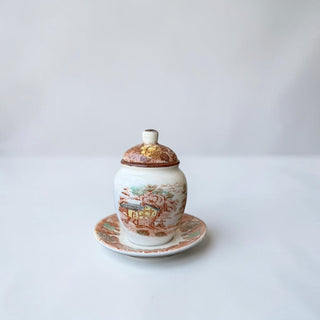 Her Good Eye vintage ceramic lidded jar with saucer, featuring a pastoral scene and floral lid. Earthy tones.