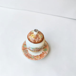 Her Good Eye vintage ceramic lidded jar with saucer, featuring a pastoral scene and floral lid. Earthy tones.