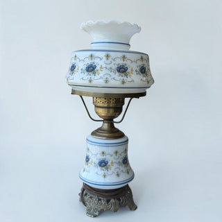 Her Good Eye vintage milk glass hurricane lamp with blue floral patterns, ruffled shade, and antique brass base.