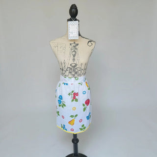 Her Good Eye vintage style white half apron with colorful fruit print, floral motifs, and yellow hem trim on a mannequin.