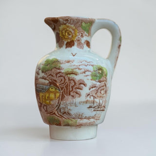 Her Good Eye vintage ceramic pitcher with pastoral landscape print, yellow house, trees, water, floral rim. Brown, green, yellow, blue.
