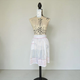 Her Good Eye white cotton half apron with red embroidery on hem and waistband, featuring a front pocket and adjustable ties.